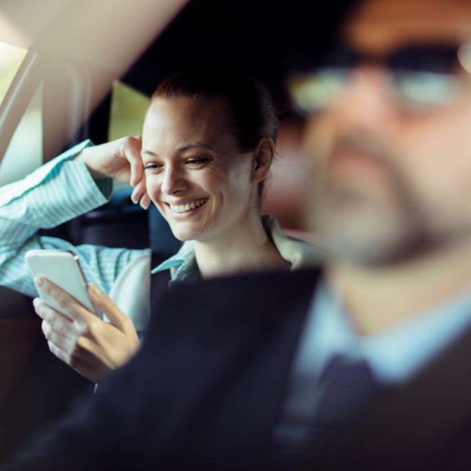 Relaxed passenger enjoying a chauffeured ride, smiling while checking her phone in the backseat, with the chauffeur in view wearing sunglasses, ensuring a smooth journey.